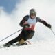 A skier carving down a snowy slope, wearing a helmet and goggles, with mountains in the background and powder flying up from their skis.